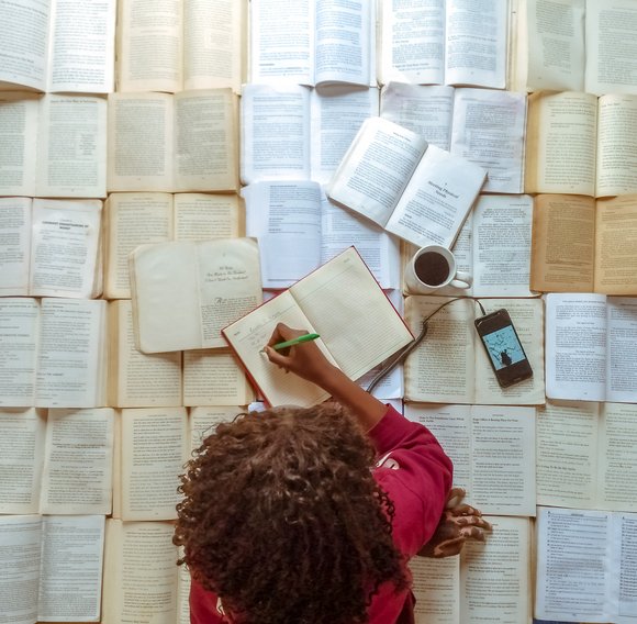 Blick von oben: Auf dem Boden ind das ganze Bild füllend Bücher aufgeschlagen. Auf den Büchern liegt eine Frau mit dunklen Locken und schreibt in ein Buch. Neben ihr stehen eine Tasse Kaffe und liegt ein Smartphone. Titel: M von Emmanuel Adegbenro.ay many books 2  