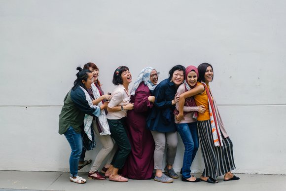 Sieben Frauen stehen in einer Reihe mit dem Rücken zum Bauch der jeweils nächsten Person. Sie stehen eng aneinander gelehnt und lachen lauthals. 