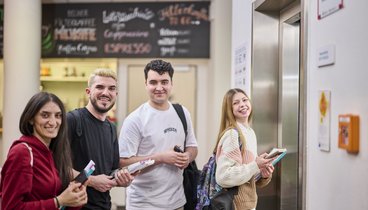 Junge Menschen warten im Erdgeschoss der VHS auf den Fahrstuhl und lachen in die Kamera