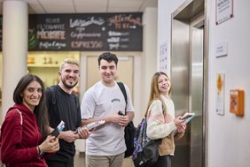 Junge Menschen warten im Erdgeschoss der VHS auf den Fahrstuhl und lachen in die Kamera