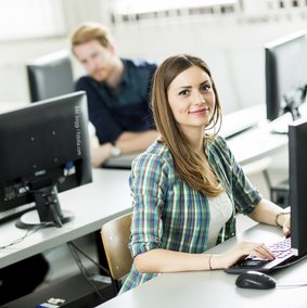 Junge Menschen sitzen in einem Seminarraum an PCs. Eine junge Frau im Vordergrund schaut in die Kamera. Bild: boggy / Fotolia.com