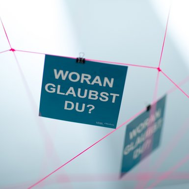 Eine quadratische Karte mit der Aufschrift „Woran glaubst du?“ hängt an einem pinken Faden. Bild: CEWE.