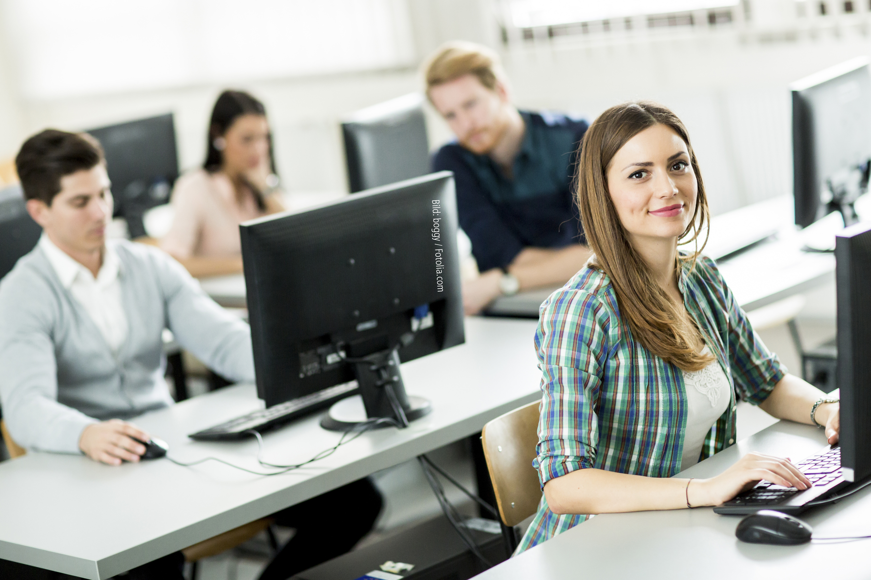 Junge Menschen sitzen in einem Seminarraum an PCs. Eine junge Frau im Vordergrund schaut in die Kamera. Bild: boggy / Fotolia.com