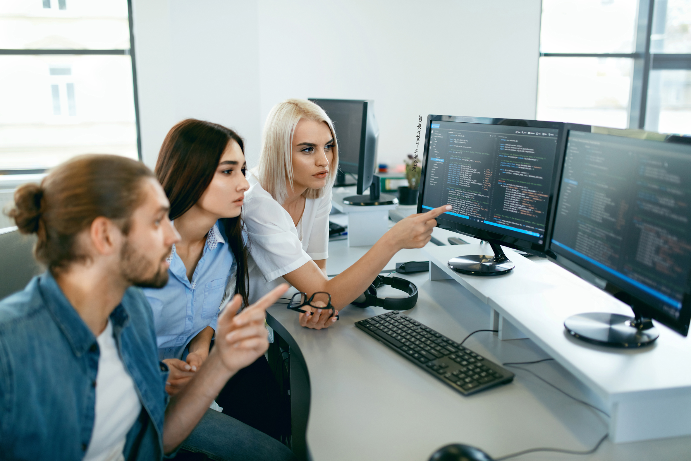 Drei junge Menschen sitzen vor einem PC mit Code und unterhalten sich. Bild: puhhha – stock.adobe.com 